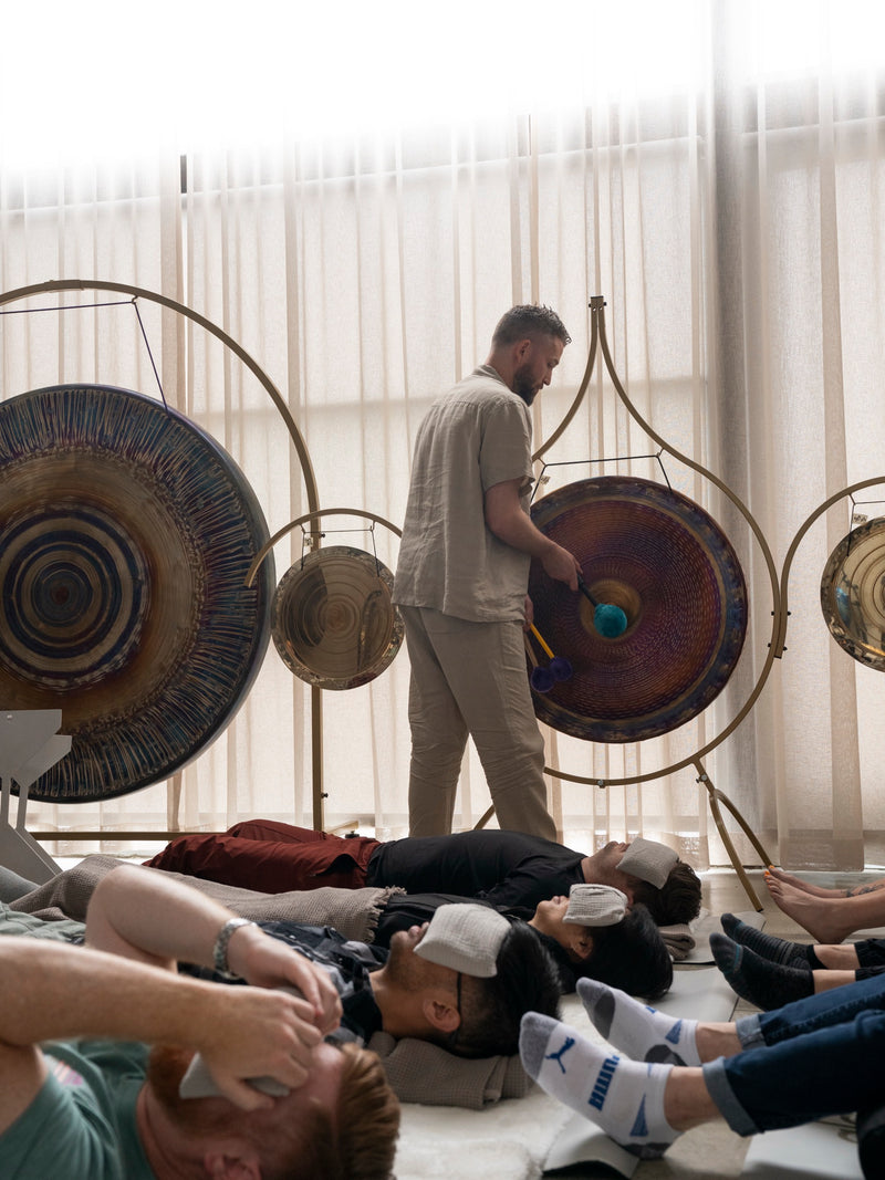Avery playing a gong in the Los Angeles studio while guests rest on mats.
